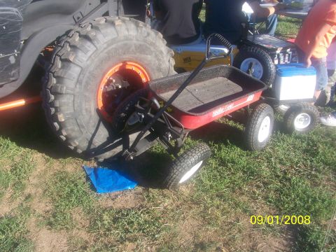 1989 Ford Bronco - A large off-road tire dwarfs a red wagon, with other vehicles visible in the background at an outdoor event.