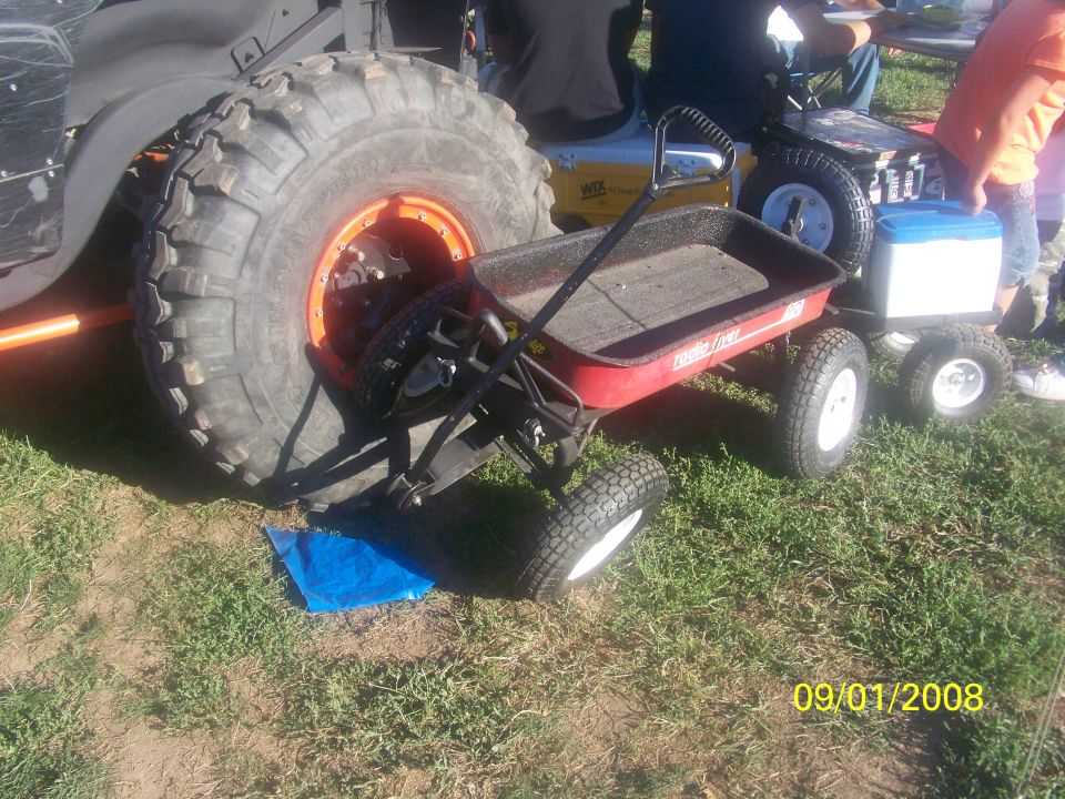 Large off-road tire next to a red Radio Flyer wagon, with other small vehicles in background