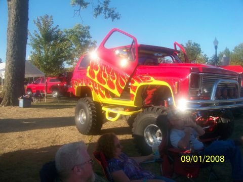 1989 Ford Bronco - A custom lifted SUV with flame graphics and open doors sits parked at an outdoor event, with another red truck visible in the background.