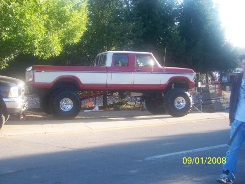 1989 Ford Bronco - A lifted red and white pickup truck with large tires is parked on the side of the road next to another vehicle.