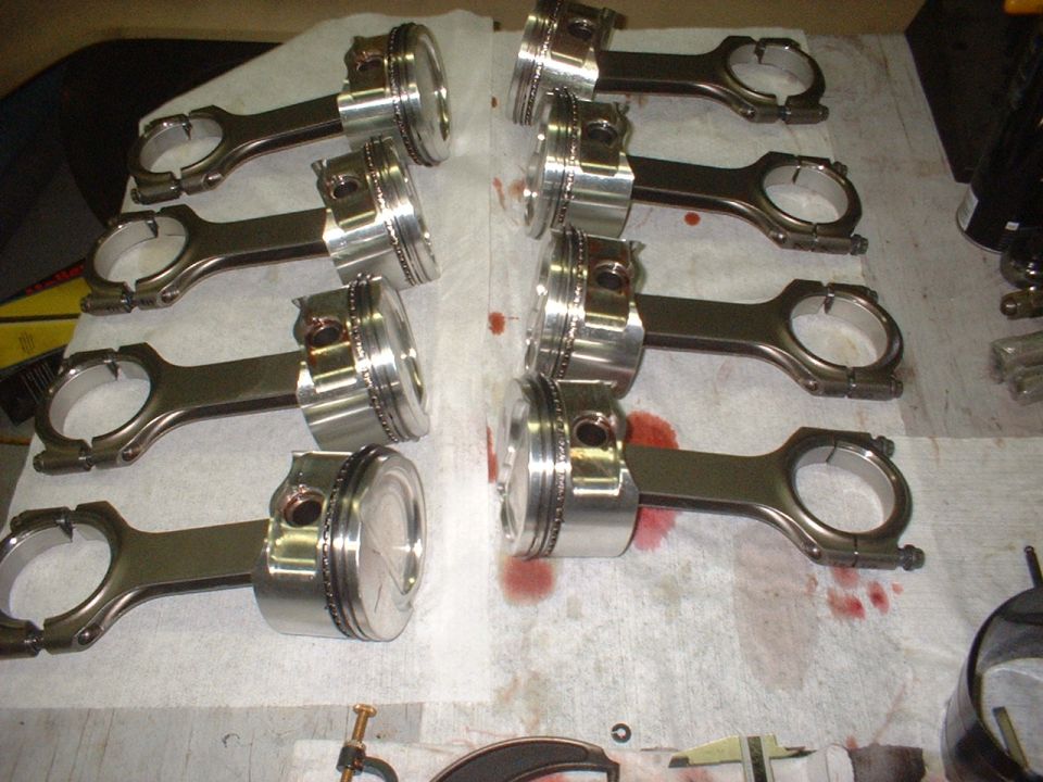 Eight pistons and connecting rods laid out on a surface with oil stains