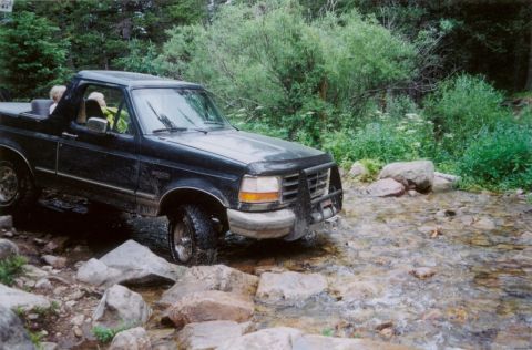 1995 Ford Bronco - A black Ford Bronco navigates a rocky stream bed surrounded by lush green foliage.