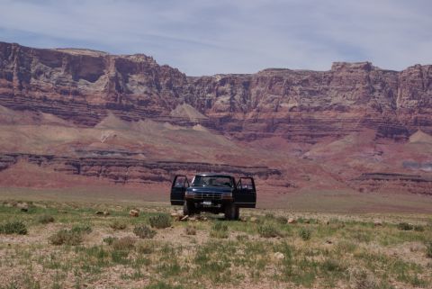 1995 Ford Bronco - A dark SUV sits in a dry, grassy field with its doors open, dwarfed by massive, layered red rock formations under a cloudy sky.