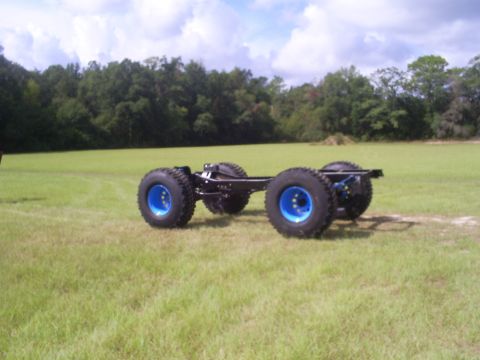 1992 Ford Bronco - This vehicle chassis, equipped with oversized tires and striking blue rims, sits on a wide expanse of grass under a partly cloudy sky.