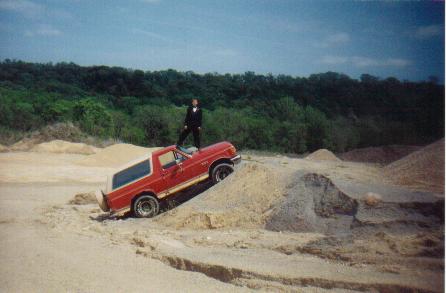 Red Ford Bronco on a dirt mound, person standing on hood. Tan top, off-road tires