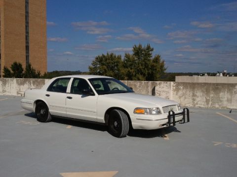1998 Ford Crown Victoria - This white sedan is parked on a concrete surface with a building and trees in the background.