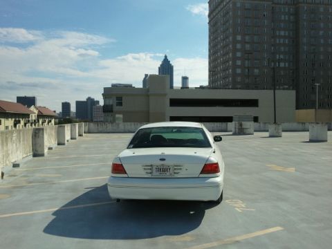 1998 Ford Crown Victoria - A white 1998 Ford Crown Victoria is seen from the rear on a rooftop parking level, with a cityscape visible behind it.