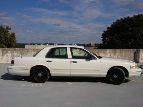 1998 Ford Crown Victoria - This white sedan is parked on a concrete surface with a wall and trees in the background under a cloudy sky.