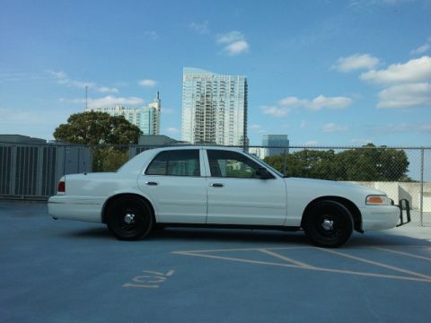 1998 Ford Crown Victoria - A white 1998 Ford Crown Victoria is parked in a lot with city buildings in the background.