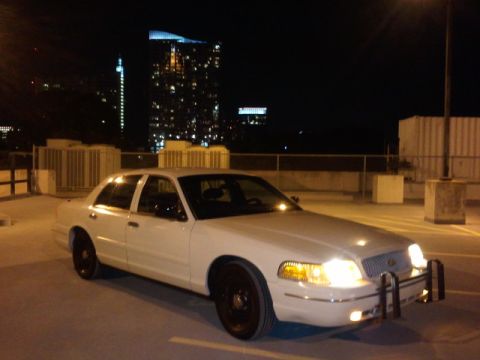 1998 Ford Crown Victoria - A white sedan with a push bumper is parked in a lot at night, with city lights in the background.