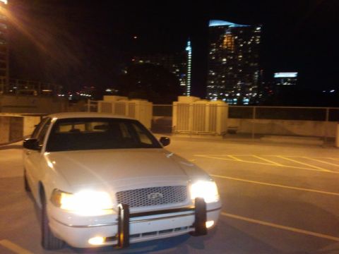 1998 Ford Crown Victoria - A white 1998 Ford Crown Victoria is parked on a rooftop at night, with the city skyline illuminated behind it.