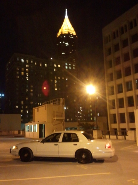 1998 Ford Crown Victoria - A white 1998 Ford Crown Victoria is parked in a lot at night, with illuminated city buildings in the background.