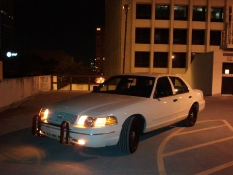 1998 Ford Crown Victoria - A white sedan with a push bumper is parked in a lot at night, with a building in the background.