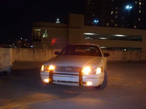 1998 Ford Crown Victoria - A white 1998 Ford Crown Victoria with a push bar is parked at night, its headlights illuminating the scene.