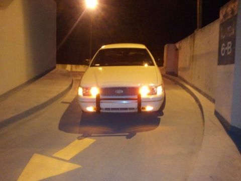 1998 Ford Crown Victoria - A white 1998 Ford Crown Victoria is parked in a dimly lit area, its headlights illuminating the road.