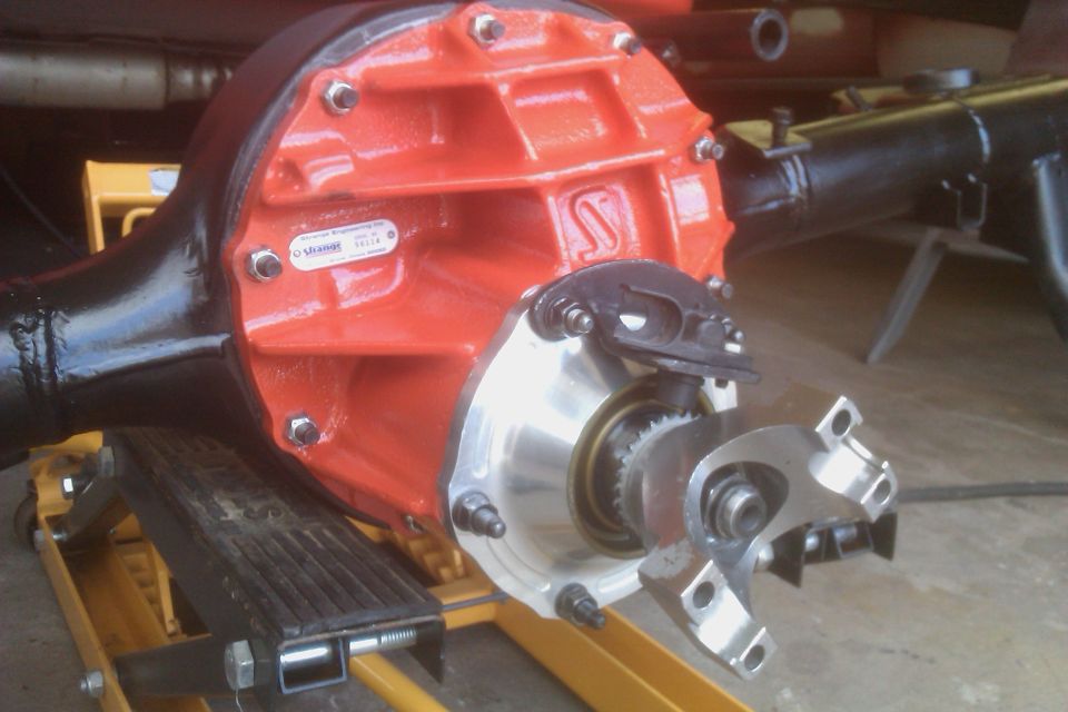 Close-up of a rear differential assembly with an orange center section and black axle tubes, supported by a yellow jack