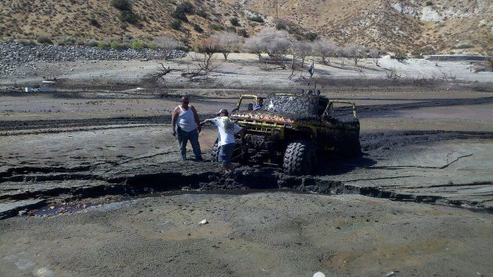 Yellow off-road vehicle covered in mud, stuck in a muddy ditch with two people standing nearby