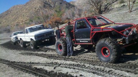 1986 Ford Bronco - A white pickup truck and a heavily modified buggy are seen on a dirt track in a desert environment.