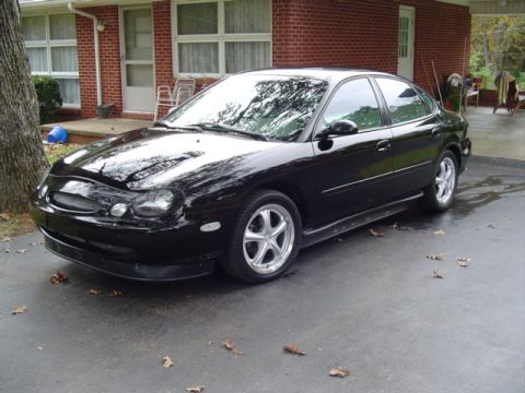 1999 Ford Taurus - Black four-door Ford Taurus sedan parked on a wet driveway in front of a brick house, shot from the front three-quarter. It wears a debadge…