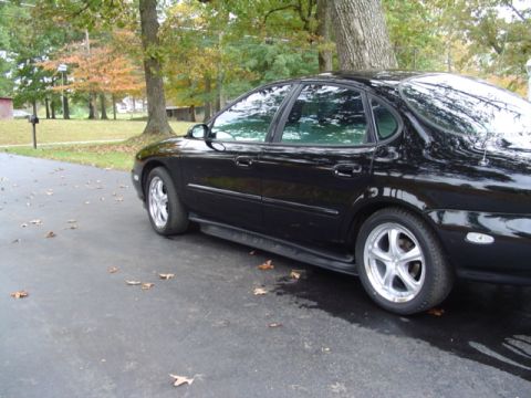 1999 Ford Taurus - Rear three-quarter view of a black Ford Taurus sedan on a driveway with autumn leaves and trees behind it. Polished five-spoke aftermarket…