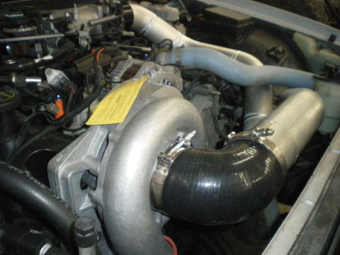 2004 Ford Crown Victoria - Close-up view of engine components, including a supercharger and custom piping, with a yellow warning tag visible.