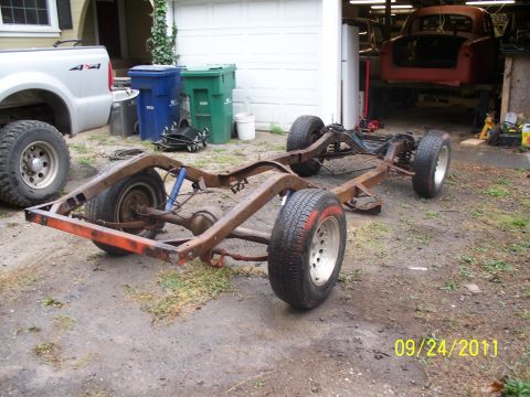 1990 Ford Bronco - A vehicle frame with wheels and suspension is shown outdoors, with a pickup truck and garage in the background.