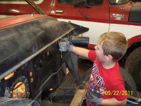 1990 Ford Bronco - A young boy works on the engine bay of a vehicle, with a red Bronco in the background.