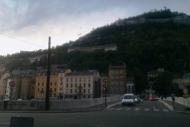 White van driving on a street with buildings and a hill in the background