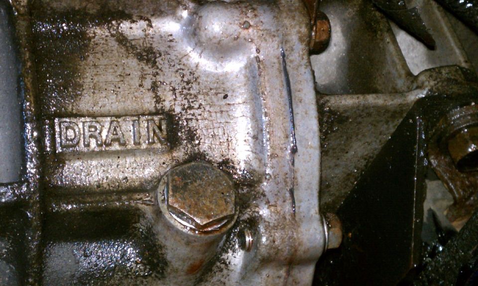 Close-up of a greasy transmission housing with the word DRAIN stamped into it and a hexagonal drain plug