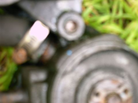 1990 Ford Bronco - A blurry shot focuses on mechanical components, with green foliage in the background and a website logo at the bottom.