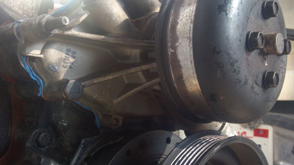 Engine water pump with blue gasket and metal pulley. Some rust and dirt visible