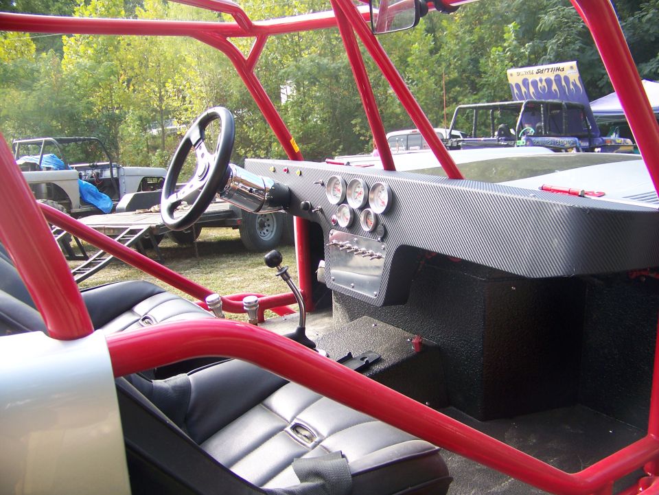Interior of a custom off-road buggy with red roll cage, black seats, and a carbon fiber dash with gauges. Another buggy and trailer are visible in the background