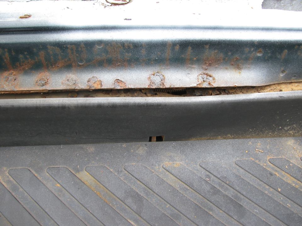Close-up of rust on a dark green vehicle panel above a textured black plastic trim piece