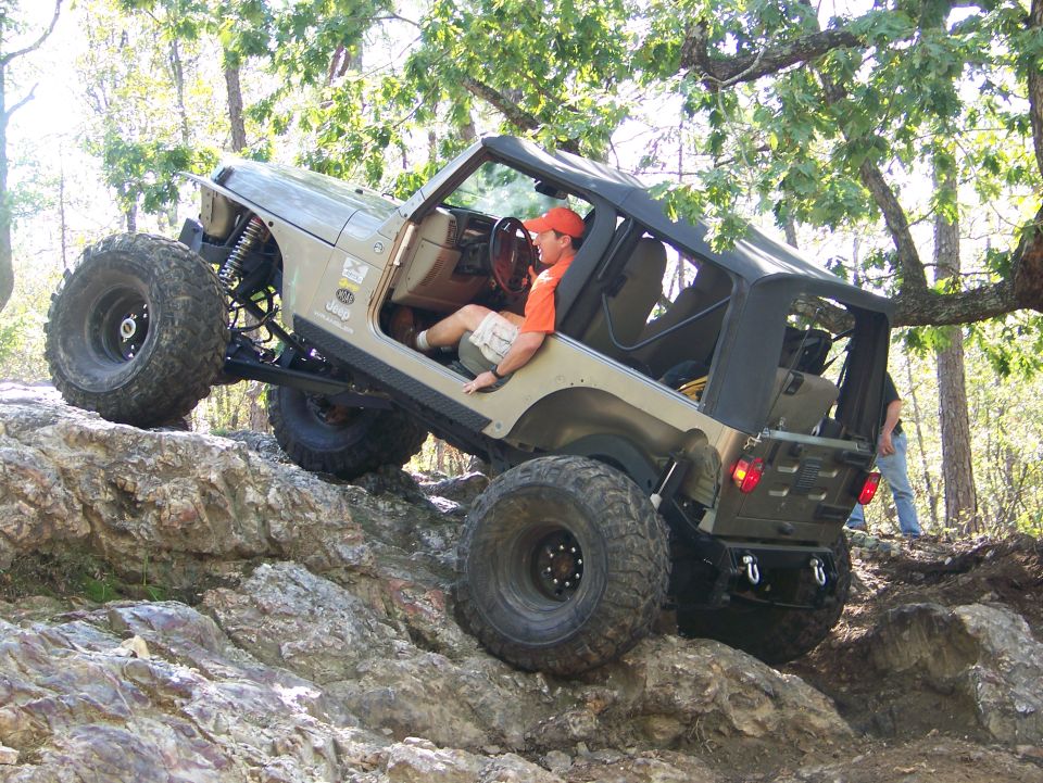 Jeep Wrangler with large tires climbing over rocks, driver wearing an orange hat