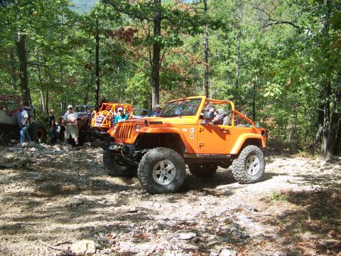 2001 Dodge 1500 - Several off-road vehicles are gathered on a wooded trail, with an orange Jeep Wrangler prominently in the foreground.