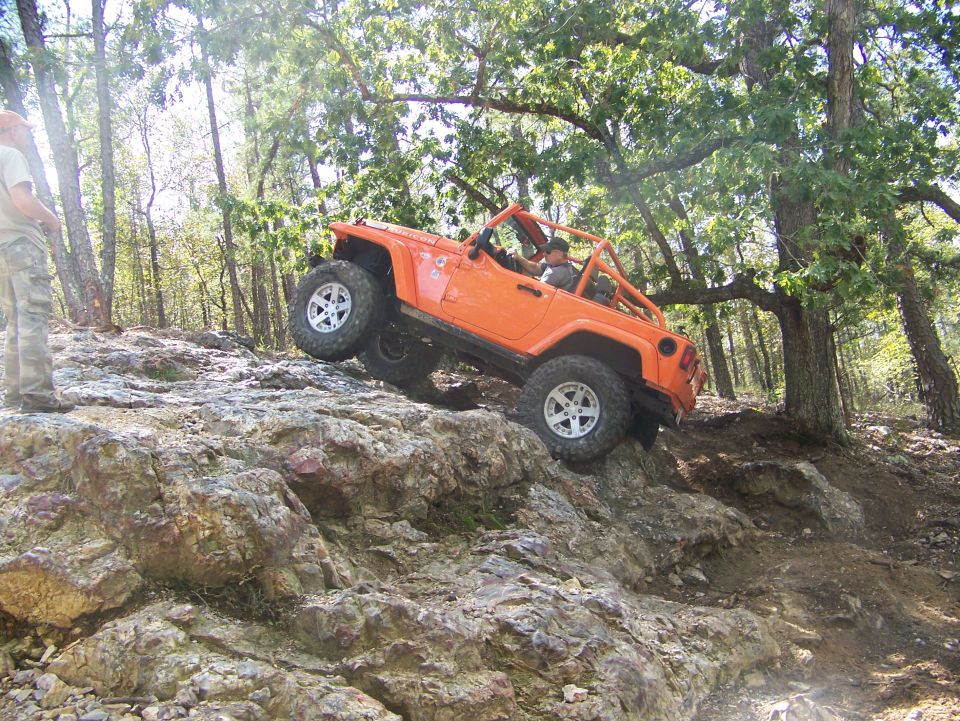 Orange Jeep Wrangler climbing a rocky hill with a person standing nearby