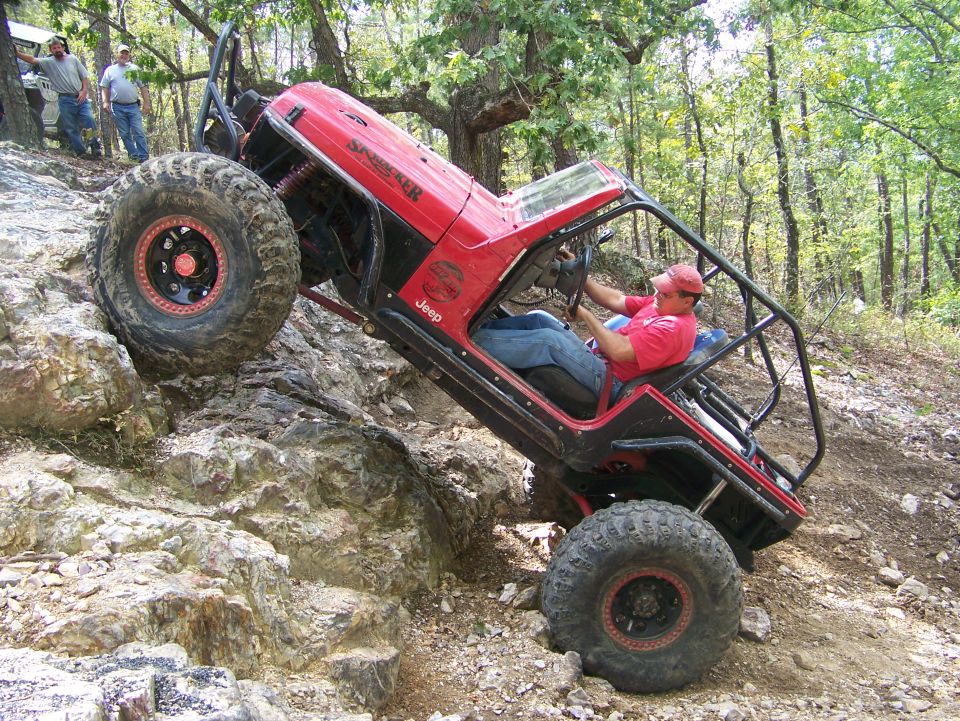 Red Jeep with roll cage climbing rocky hill, driver wearing red hat and shirt