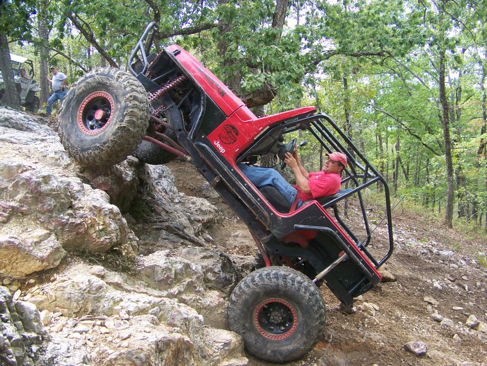 Red Jeep climbing a rocky hill with a person driving. Another off-road vehicle is visible in the background
