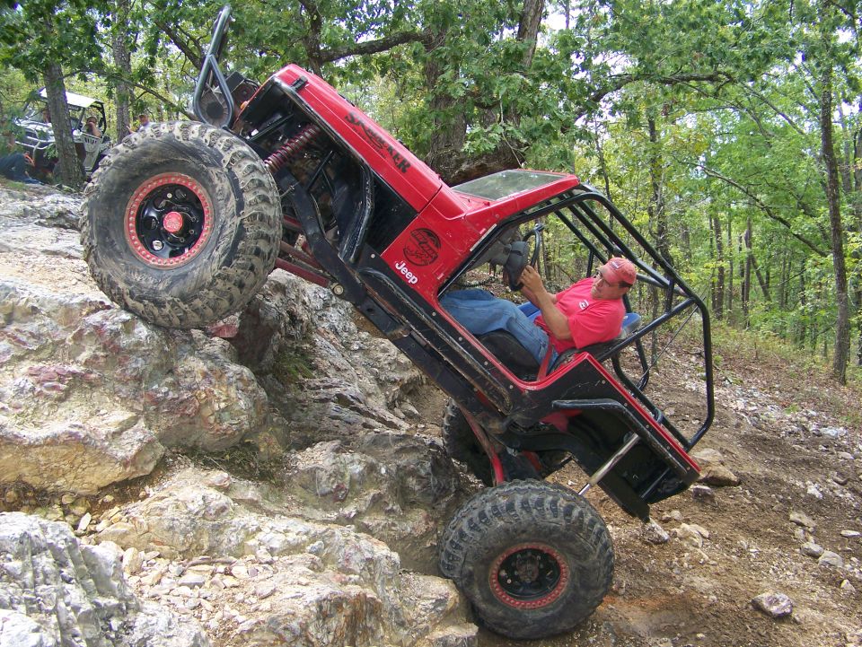 Red Jeep climbing rocks with a driver inside, another vehicle visible in background