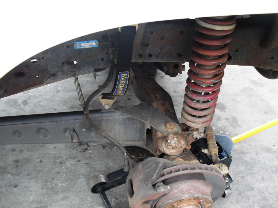 Vehicle suspension with red coil spring, rusty brake rotor, and a black strap labeled "McKenzie"