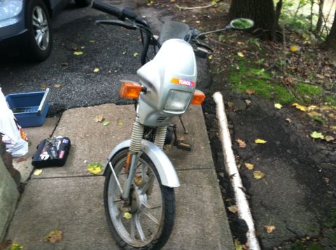 1999 Tomos Targa - A silver Tomos moped is parked on a concrete path next to an asphalt driveway, with a car in the background.