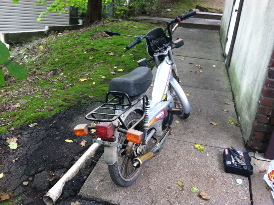 Silver moped with black seat and rack, rear view. Text on side reads 'Tomos'