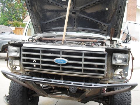 1986 Ford Bronco - Front view of a modified Ford Bronco, showing its grille and open hood. Another vehicle is partially visible to the left.