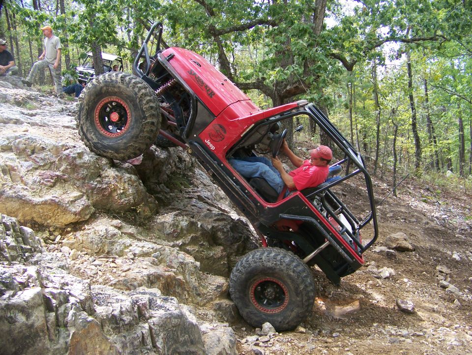 Red Jeep Wrangler with roll cage climbing a steep, rocky hill. Driver in red shirt. Other people watch