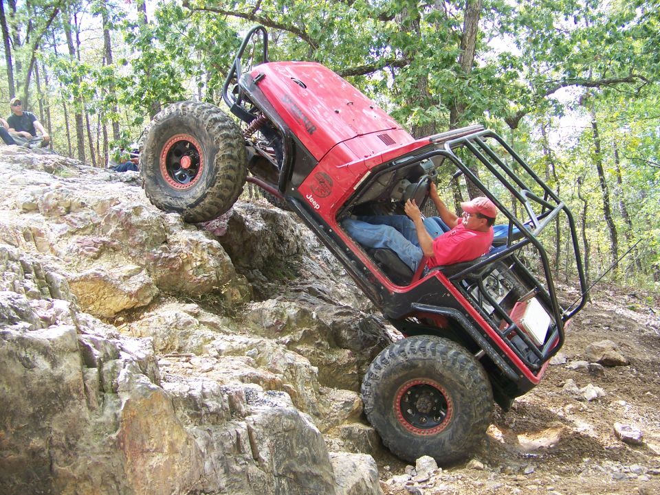 Red Jeep Wrangler with roll cage climbing a rocky incline, driver wearing a red hat