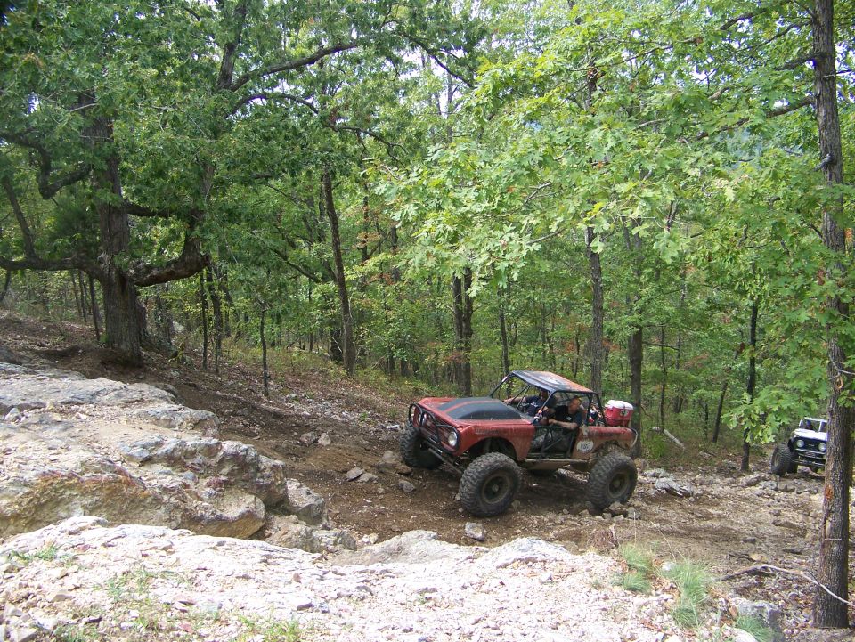 Red off-road vehicle with large tires on a rocky trail, another white vehicle behind it