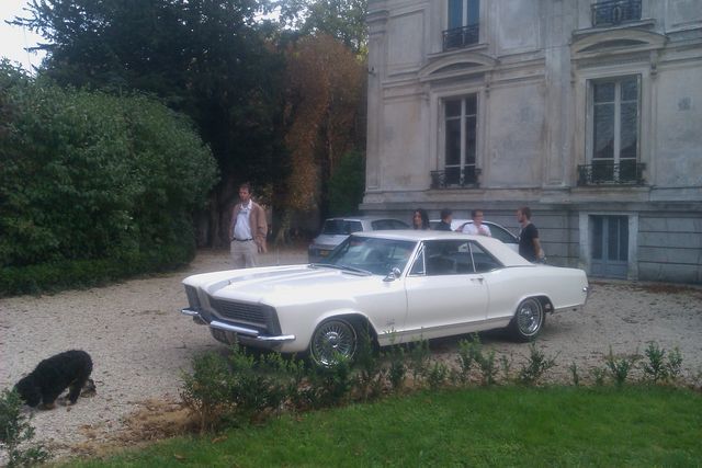 White coupe parked in front of a large building with a man standing nearby and a dog on the left