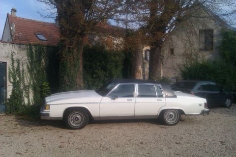 1991 Ford LTD Crown Victoria - A white four-door sedan with a black vinyl roof is parked on a gravel surface in front of a building.