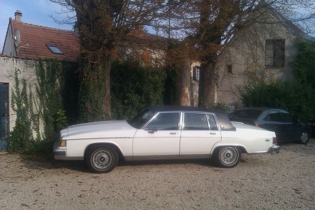 White four-door sedan with a black vinyl roof, parked on gravel. A dark sedan is visible behind it
