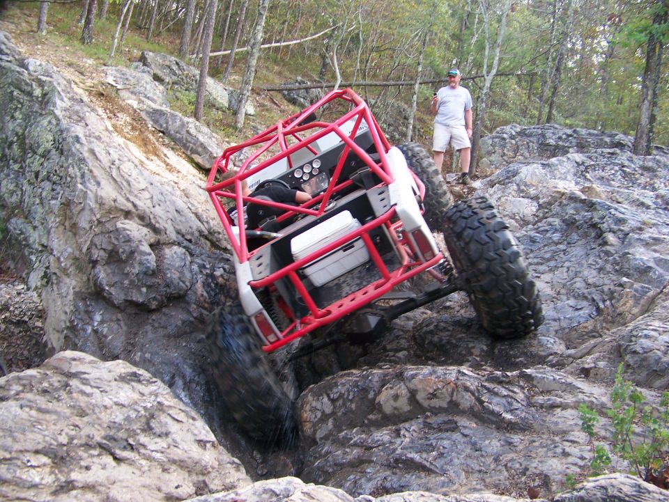 Off-road buggy with a red roll cage going over rocks, a man stands nearby